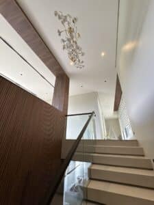 A modern staircase with glass railing leads to an upper floor. Wood paneling on the left wall, a decorative chandelier hangs from the ceiling, and natural light fills the space.