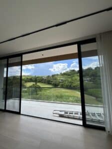 A glass sliding door opens to a balcony with a glass railing, overlooking a green landscape and partially cloudy sky. A ladder lies on the balcony floor.