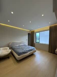 Minimalist bedroom with a large bed and chair covered in plastic, wooden floor, recessed ceiling lights, and a window with beige curtains overlooking greenery.