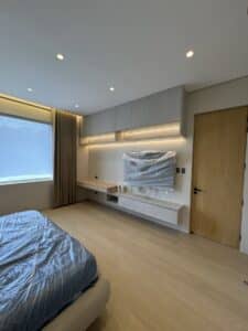 Modern bedroom with a neatly made bed, large window with curtains, built-in desk and shelves, and a wall-mounted TV covered in plastic. Light wood flooring and recessed ceiling lights.
