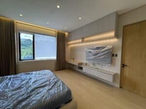 Modern bedroom with a large window, a bed and furniture covered in plastic, built-in desk and cabinets, and wood accents, suggesting the room is newly finished or being prepared.