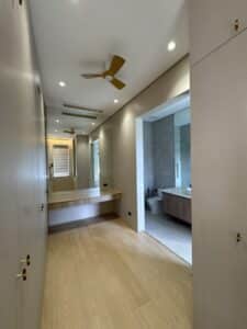 A narrow dressing room with built-in wardrobes, a wooden vanity, ceiling fans, large mirrors, and an open doorway leading to a modern bathroom.