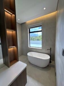 Modern bathroom with a freestanding oval bathtub, floor-mounted black faucet, large window, built-in shelving, and neutral-toned tiles. Natural light fills the room.