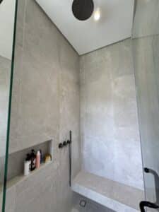 Modern walk-in shower with gray stone tiles, built-in shelf with toiletries, black rainfall showerhead, handheld shower, and a bench along the wall.