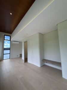 Minimalist, empty room with light wood floor, white walls, a built-in wooden bench, large window, and ceiling lights.