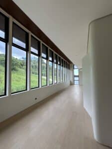 A hallway with large floor-to-ceiling windows on one side, light wood flooring, and white walls, overlooking green grassy hills and trees outside.