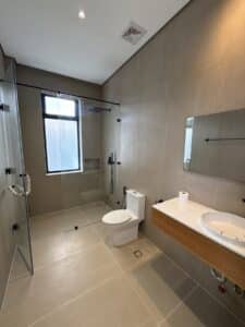 Modern bathroom with beige tiles, glass-enclosed shower, wall-mounted sink with mirror, and a toilet. Natural light comes in through a frosted window.
