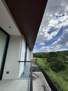 View from a modern balcony with glass railing, overlooking green landscape and cloudy blue sky; construction materials are visible on the ground below.