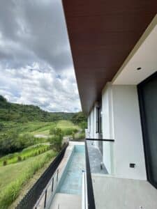 Modern house with a balcony overlooking a narrow, empty pool and a green landscape with hills under a cloudy sky.