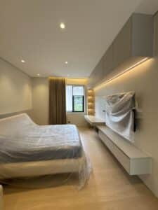 Modern bedroom with light wood floors and built-in shelves; bed and TV are covered in protective plastic, and natural light enters through a window with tan curtains.
