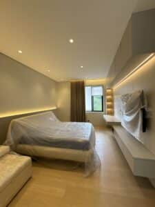 Modern bedroom with a bed, sofa, and wall-mounted TV, all covered in protective plastic, featuring wood flooring, built-in shelves, and a window with closed curtains.