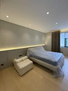 A modern bedroom with a large bed and ottoman covered in plastic, featuring recessed lighting, beige walls, wood flooring, and a window with brown curtains.