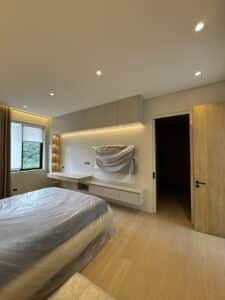 Minimalist bedroom with wooden floor, a bed covered in plastic, built-in cabinets and desk, wall shelves with lights, a window with curtains, and an open wooden door.