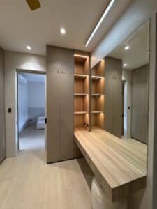 Modern walk-in closet with built-in beige wardrobes, open wooden shelves with lights, a wooden vanity table, and a large wall mirror. Bedroom visible through open doorway.