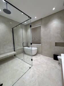 Modern bathroom with a glass-enclosed shower, a freestanding bathtub near a window with a closed blind, and beige tiled walls and floors.