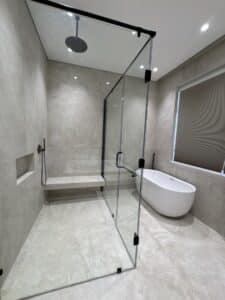 Modern bathroom with glass shower enclosure, built-in bench, overhead rain shower, freestanding bathtub, beige stone tiles, and a window with a closed blind.