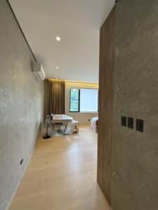 A modern, minimally furnished room with a covered table, floor lamp, wooden floor, air conditioner, and large window with curtains overlooking greenery.