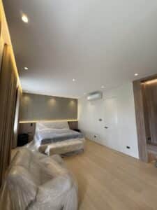 A modern bedroom with a bed and couch covered in plastic, wood flooring, recessed lighting, and neutral-colored walls.