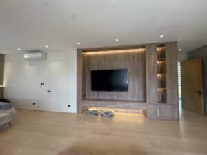 A modern living room with a large wall-mounted TV on a wooden panel, built-in shelves, two cushions on the floor, and minimal furniture.