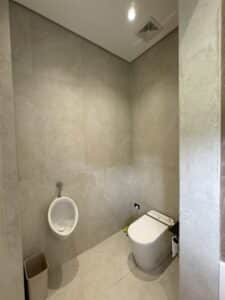 A restroom with beige tiled walls featuring a urinal, a toilet with a bidet, a small trash bin, and a roll of toilet paper on the floor.
